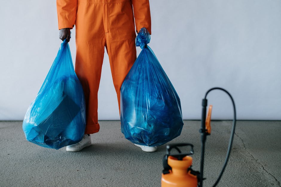 A person standing outdoors on a grassy area, wearing a plaid shirt, a green jacket, and green gloves, is holding open a large black trash bag with both hands. The bag appears to be made of thick plast