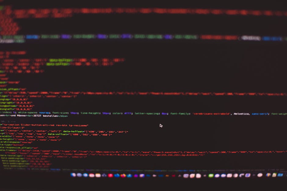 A man with glasses and a beard is seated at a wooden desk working on a computer with dual monitors. The monitors display lines of code and text files in a dark-themed interface, with one screen showin